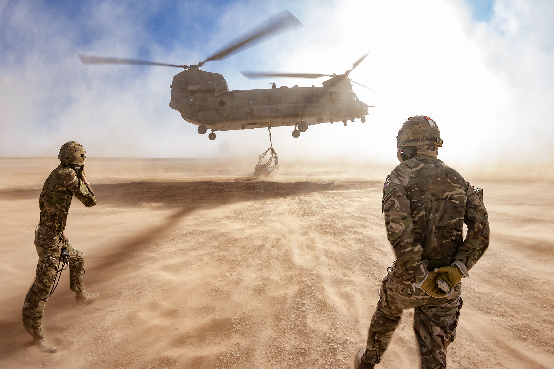 Image shows aviators looking towards helicopter carrying under-slung load in the desert.